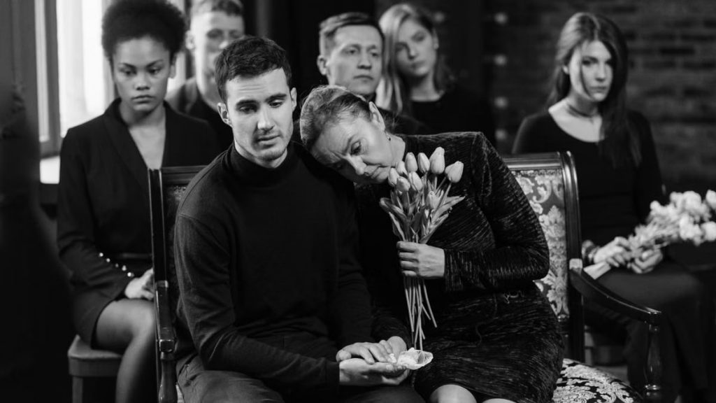 A grayscale scene of individuals gathered indoors, expressing sorrow and loss at a funeral.